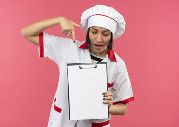 厨师印象深刻的年轻女厨师在厨师制服举行看 并指着剪贴板上的粉红色与复制空间隔离烹饪女年轻