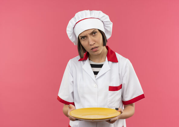 粉困惑的年轻女厨师穿着厨师制服拿着空盘子孤立在粉红色的复制空间年轻空制服