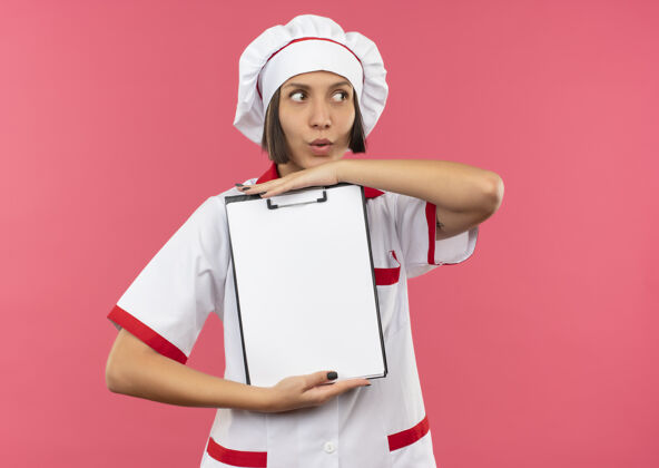 女印象深刻的年轻女厨师穿着厨师制服拿着剪贴板 看着旁边孤立的粉红色年轻举行烹饪