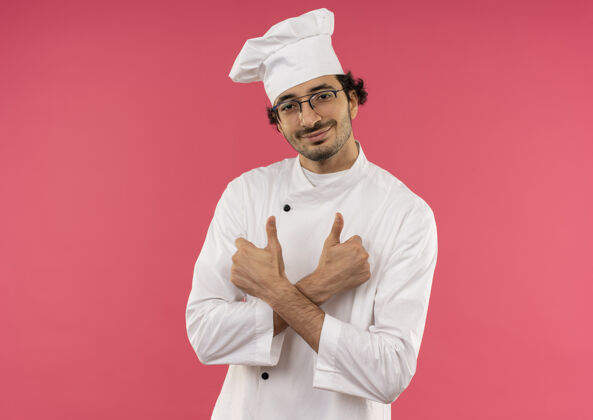 年轻面带微笑的年轻男厨师穿着厨师制服 戴着眼镜双手交叉 他的大拇指孤立地竖立在粉红色的墙上眼镜微笑拇指