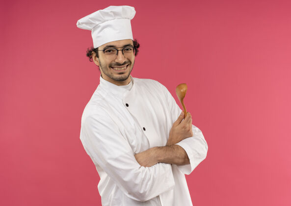 拿着面带微笑的年轻男厨师穿着厨师制服 戴着眼镜 拿着勺子 双手交叉 隔离在粉红色的墙上 留着复印空间制服穿微笑
