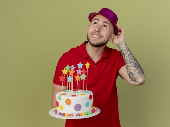派对微笑的年轻帅哥斯拉夫党人戴着党的帽子触摸帽子举行生日蛋糕看着孤立的橄榄绿背景与复制空间抱着蛋糕微笑
