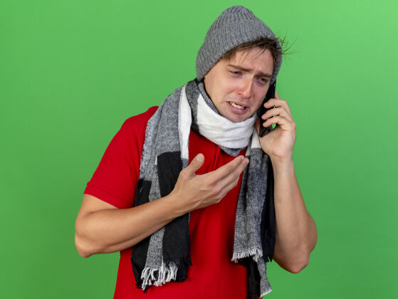 英俊悲伤的年轻英俊的金发病男子戴着冬天的帽子和围巾打电话看着一边保持手在空气中隔离在绿色背景与复制空间年轻壁板电话