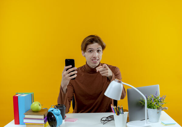 表演高兴的年轻学生男孩坐在书桌旁 手里拿着学习工具 拿着电话 在黄色的墙上向你们展示孤立的手势男孩坐着书桌