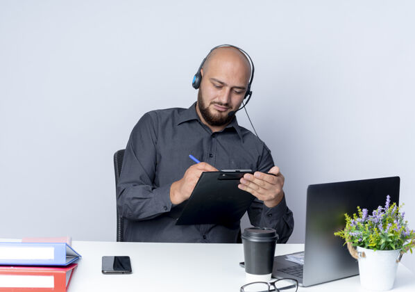 男人专注的年轻秃头呼叫中心男子戴着耳机坐在办公桌旁 用写字笔在写字板上写字 孤立在白纸上工作电话工具