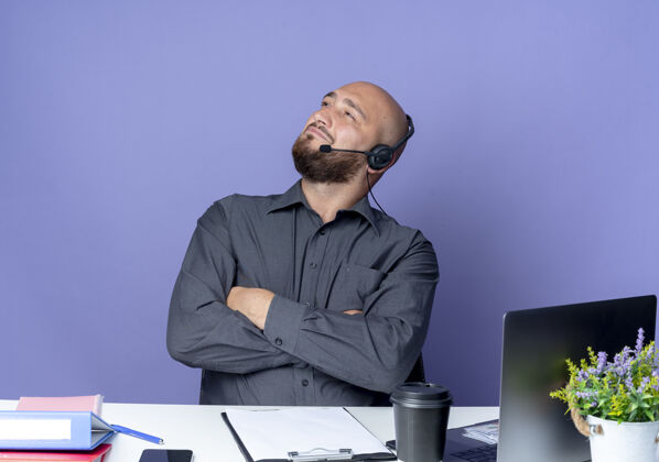 紫色自信的年轻秃头呼叫中心男子戴着耳机坐在办公桌前 紧闭的姿势 带着工作工具 孤立地仰望紫色办公桌年轻电话