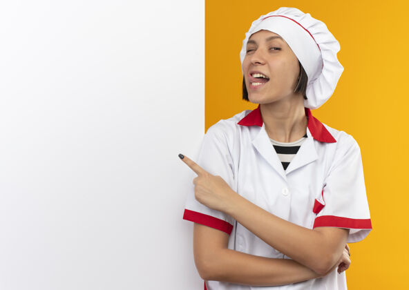 空间自信的年轻女厨师身着厨师制服 站在白色墙壁前 露出舌头 指着隔离在橙色墙上的墙壁 留有复制空间信心复制烹饪