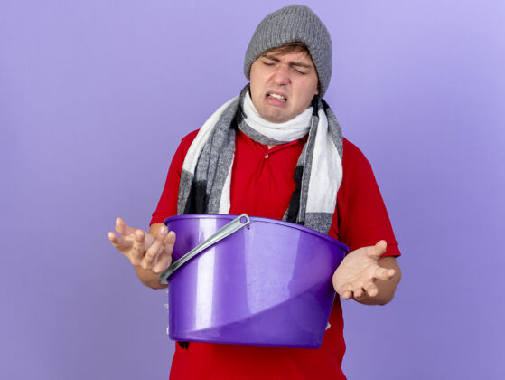 围巾恼怒的年轻英俊的金发病男子戴着冬天的帽子和围巾拿着塑料桶闭着眼睛隔离在紫色背景与复制空间疾病恼火年轻