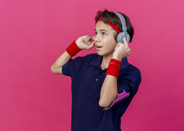 袖带令人印象深刻的年轻英俊的运动男孩戴着头带和腕带和耳机手机袖带与牙套看直触摸耳机在深红色背景与复制空间隔离牙科年轻电话
