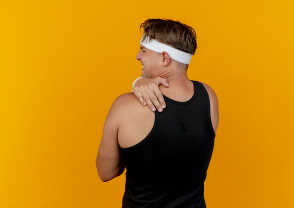 橙色疼痛的年轻英俊的运动男子戴着头带和腕带站在后面的看法把手放在后面孤立的橙色与复制空间疼痛站立佩戴