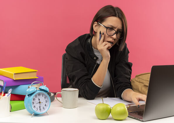 学生专注的年轻女学生戴着眼镜坐在书桌旁拿着笔把手放在脸上用笔记本电脑隔离在粉色上笔记本电脑拿着粉色