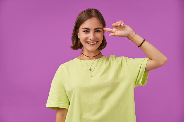 20多岁十几岁的女孩 快乐快乐 深色短发手指靠近太阳穴站在紫色的墙上穿着绿色t恤 牙套 项链 手镯和戒指项链表演休闲