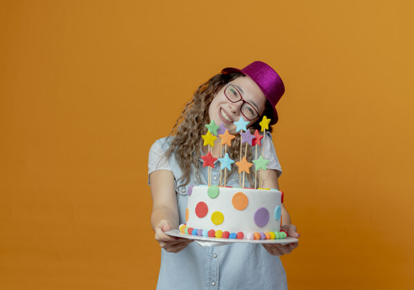 微笑歪着头微笑的年轻女孩戴着眼镜和粉红色的帽子拿着生日蛋糕孤立在橙色的墙上抱着女孩倾斜