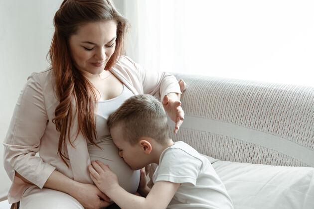 人小男孩在家里的沙发上拥抱和亲吻怀孕母亲的肚子房子女人窗户