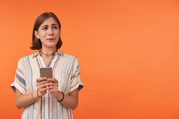 不安照片中的可爱女士手持智能手机 在橘色墙上的复印区右侧观看 展示她的担忧穿着条纹衬衫 项链和手镯聪明信息女性