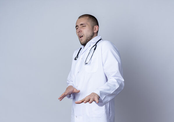 视图恼怒的年轻男医生穿着医用长袍 脖子上戴着听诊器 站在侧视图中 伸出双手隔离在白色的空间里年轻人周围空间