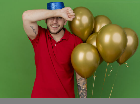 气球面带微笑的年轻英俊的斯拉夫党人戴着党的帽子拿着气球保持手臂在前额前看着隔离在绿色背景上的相机额头派对帽子