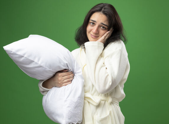 女孩高兴的年轻白种人生病的女孩穿着长袍抱着枕头手放在脸上看着隔离在绿色背景下的相机相机抱着脸