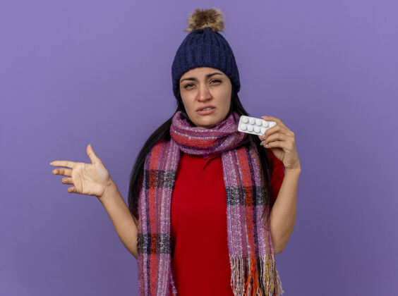 壁板虚弱的年轻白种人生病的女孩 戴着冬天的帽子 戴着围巾 展示着一包平板电脑 看着摄像机指着一边 手被隔离在紫色背景上 留着复印空间手疾病弱