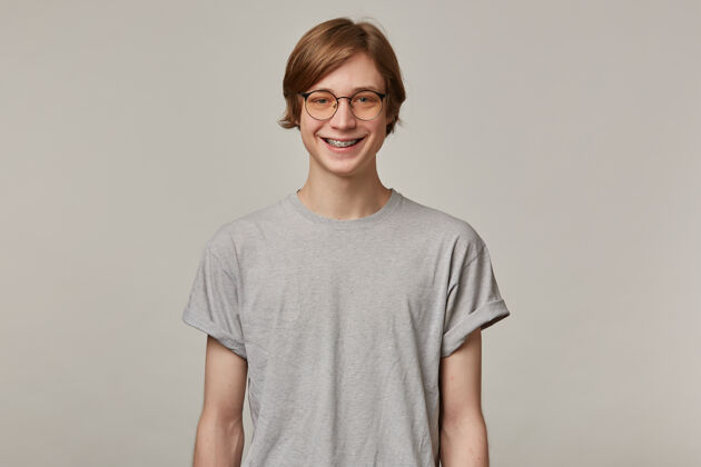 青少年快乐的 金发的成年男性的肖像穿着灰色t恤 戴着眼镜 戴着牙套人和情感的概念微笑隔着灰色的墙欢呼金发肖像