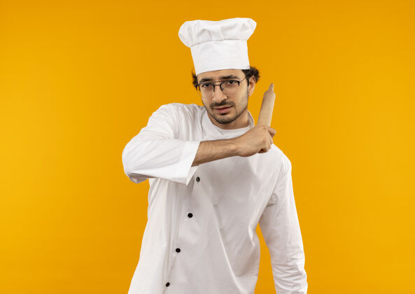 抱年轻的男厨师穿着厨师制服 戴着眼镜 肩上扛着擀面杖 隔离在黄色的墙上制服年轻绕