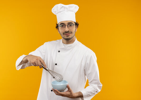厨师年轻的男厨师穿着厨师制服 戴着玻璃杯 手里拿着威士忌和碗 隔离在黄色的墙上打蛋请制服