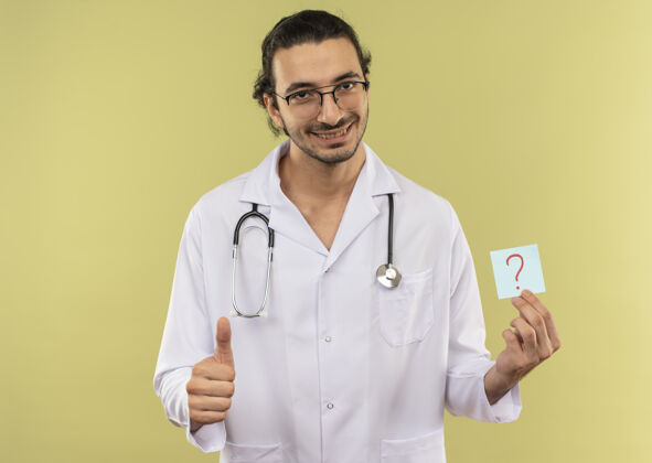 微笑微笑的年轻男医生 戴着眼镜 身穿白色长袍 手持听诊器 拿着纸问号 他的大拇指竖立在隔离的绿色墙壁上 留着复印空间长袍听诊器年轻