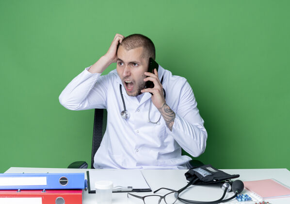 医生愤怒的年轻男医生穿着医用长袍和听诊器坐在办公桌旁 拿着工作工具讲电话 手放在头上 看着绿色隔离的一面长袍电话年轻