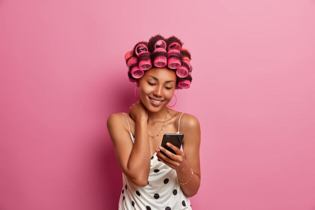 高兴快乐的黑皮肤成年女子 带着卷发器 在特殊场合做发型 穿着连衣裙 拿着手机 上网看新闻 滚动新闻 女人头发手机
