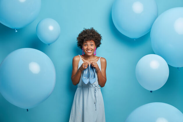 乐观快乐快乐的年轻女性穿着蓝色的连衣裙 时尚的鞋子 选择一种颜色的服装和鞋子 去庆祝生日 摆姿势人卷发请