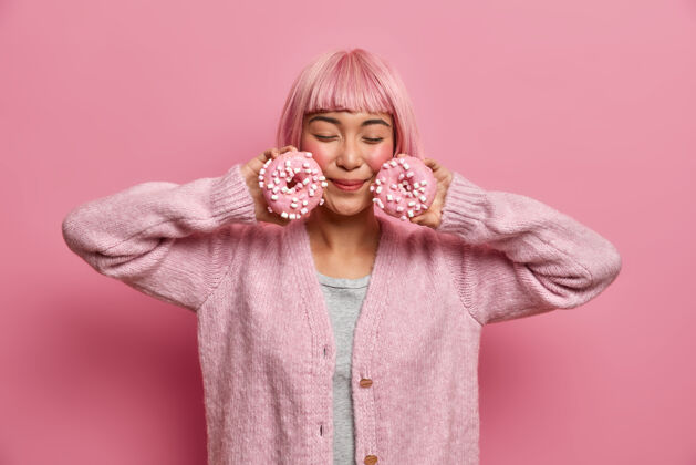 垃圾年轻的粉红头发女人喜欢美味的甜甜圈 闭着眼睛摆姿势 把甜甜圈撒在脸上 穿着暖和的毛衣 女性味道粉红头发