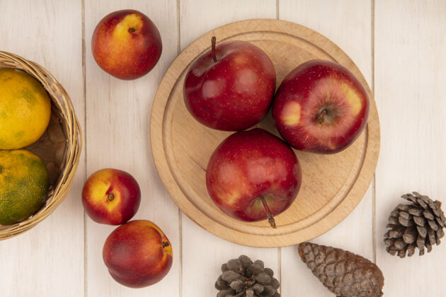 木材木制厨房板上的多汁红苹果俯视图 桶上放着橘子 白色木质墙壁上隔离着桃子和松果等级苹果视图