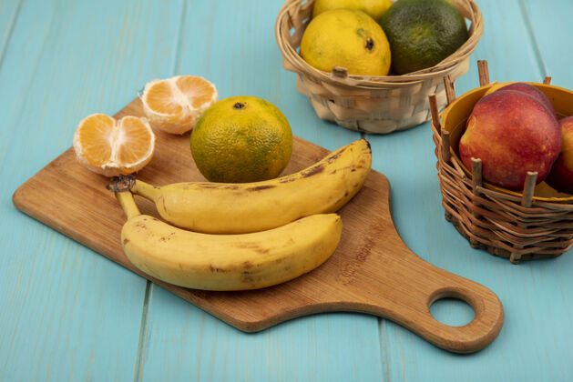 桃子木制厨房板上的多汁的整个和半个橘子的俯视图 蓝色木制墙上的桶上放着香蕉和桃子橘子香蕉新鲜