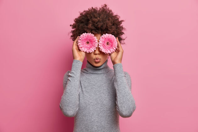 玩浪漫美丽的卷发女人把粉色非洲菊戴在眼睛上 有春天的心情 穿着休闲的灰色高领毛衣 准备用鲜花做花束灰色安排卷曲