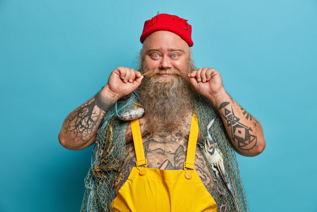 男人高兴的渔夫卷起胡子 留着浓密的胡须 肩上扛着渔网 闲暇时为了爱好和灵魂 戴着红帽子和工作服 衣衫褴褛的身体高兴红色大