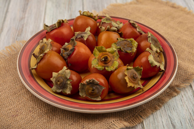 木头新鲜多汁的柿子放在一个盘子里 在灰色的木墙上的布袋上新鲜食物柿子