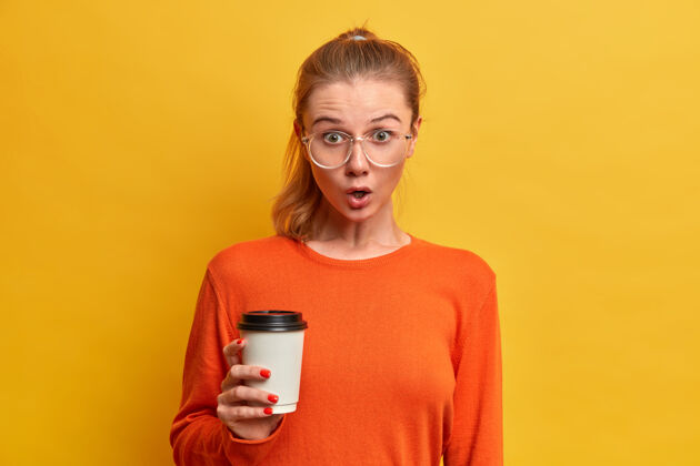 年轻人情绪恍惚的女生有咖啡休息时间 拿着一次性卡布奇诺杯 戴着大透明眼镜 穿橘黄色毛衣 听到关于团友的新鲜八卦 喝着饮料印象瞳孔