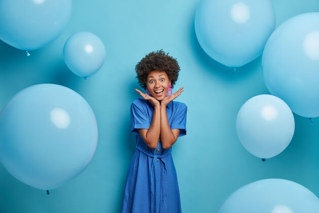 姿势笑容可掬的美国黑人女孩将手掌放在脸上 享受美妙的夏日派对 身着蓝色长裙在充气气球上摆姿势 心情愉快庆祝和生活理念充气手掌年轻