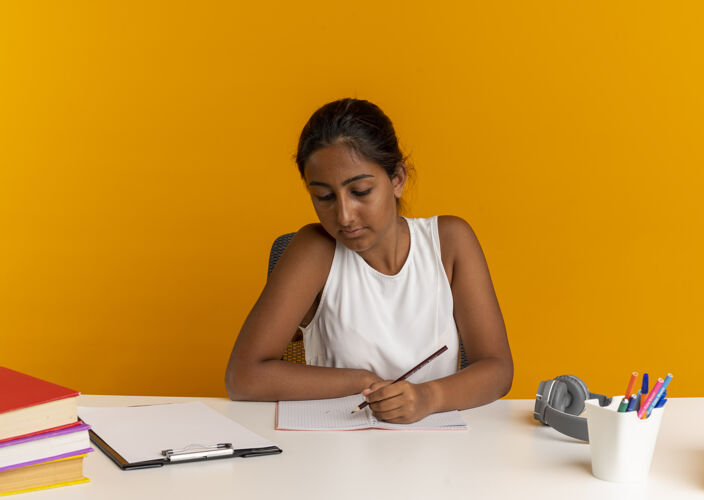 学校坐在书桌旁拿着学习用具的年轻女学生在桔子上的笔记本上写些什么年轻书桌坐着