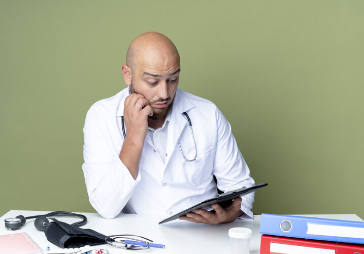 持有惊讶的年轻秃头男医生穿着医用长袍和听诊器坐在办公桌上工作听诊器绿色医疗