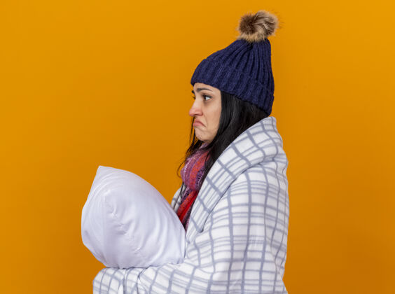 女孩困惑的年轻白种人生病的女孩戴着冬天的帽子和围巾裹着格子呢站在侧视图抱着枕头看直孤立的橙色背景与复制空间疾病格子帽子