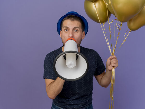 英俊戴着蓝色帽子的帅哥拿着氦气球对着紫色墙壁上孤立的扩音器说话人人人