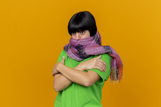 手臂冰冷的生病的年轻妇女戴着围巾 用围巾捂住嘴 双手交叉放在胳膊上 看起来像是被隔离在橙色的墙上人交叉围巾