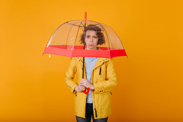 担心雨伞下站着一个愁眉苦脸的短发美女穿着雨衣 手持时尚阳伞 心烦意乱的高加索妇女的画像优雅肖像雨