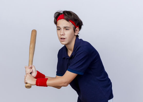 壁板令人印象深刻的年轻英俊的运动男孩戴着头带和戴着牙套的腕带看着一边拿着棒球棒打棒球在白色背景上与复制空间隔离举行玩白色