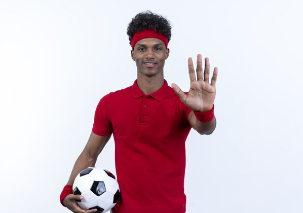 年轻微笑着的年轻的非裔美国人运动男子戴着头带和手环拿着球 显示停止手势隔离在白色表情运动停止