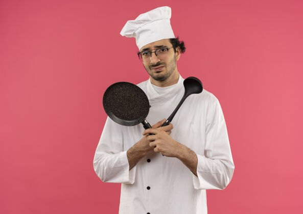 男性年轻的男厨师穿着厨师制服 戴着眼镜 手里拿着平底锅 手里拿着抹刀十字眼镜手持