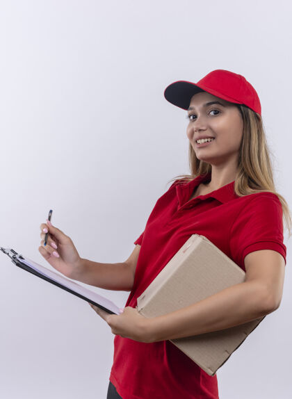 制服微笑着的年轻送货女孩穿着红色制服 戴着帽子 拿着盒子 用白纸上孤立的笔在剪贴板上写字剪贴板快递年轻人