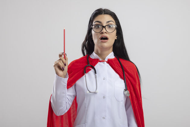 听诊器令人印象深刻的年轻白种人超级英雄女孩戴着眼镜和听诊器拿着红铅笔看着相机隔离在白色背景与复制空间年轻眼镜复制