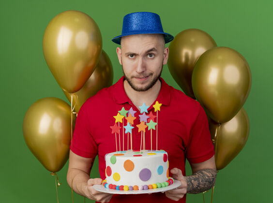 年轻可疑的年轻帅哥戴着派对帽站在气球前拿着生日蛋糕看着前面隔离的绿色墙壁市民前面怀疑